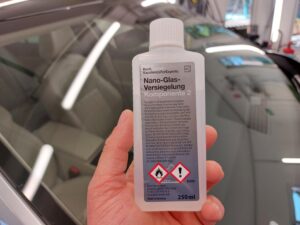 Hand holding a 250 ml bottle of Koch-Chemie Nano-Glass-Versiegelung Component 2 in front of a car windshield; hazard symbols and German warnings visible.