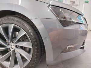 Close-up of a gray car's front left corner showing the headlight, fender, and large alloy wheel with tire.